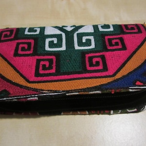 May include: A rectangular wallet with a vibrant geometric pattern in pink, green, white, orange, and blue. It features a black zip and a patterned wrist strap. The design is reminiscent of traditional textiles.
