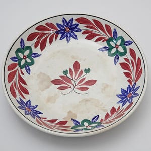 Op de afbeelding: Een ronde, witte keramische schaal met een zwarte rand. De schaal is versierd met een bloemmotief in rood, blauw en groen. Het ontwerp omvat gestileerde bloemen en bladeren, die een decoratieve rand vormen.