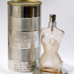Puede incluir: Un frasco de perfume con forma de torso femenino, con un pulverizador plateado, se encuentra junto a un recipiente plateado con forma de lata. El recipiente tiene grabado "Jean Paul Gaultier". El frasco contiene 50 ml.