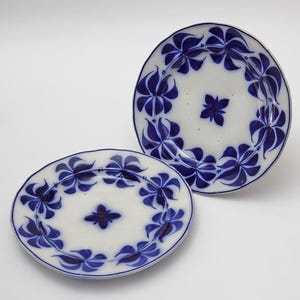 May include: Two white ceramic plates with a blue floral design. The plates have a central floral motif and a border of stylized blue flowers. The plates are oval and round, with a slightly irregular edge.