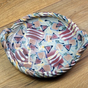 May include: A ceramic serving tray with a braided rim and handles. The tray's interior features a colourful abstract design with geometric shapes in shades of red, blue, and yellow. The background is a light blue.