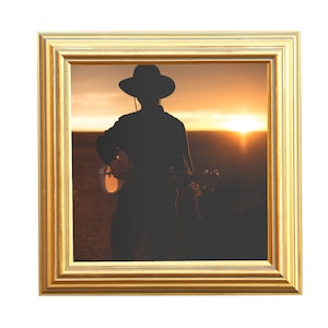 May include: A framed photograph featuring a silhouette of a person playing a banjo at sunset. The person is wearing a hat and dark clothing. The frame is gold-colored with a detailed, layered design.