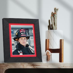 May include: Black framed photo of a firefighter wearing a helmet with the number 417. The photo has a red matting. A cactus in a white pot with a wooden stand is to the right.