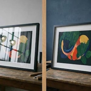 May include: Two framed abstract art prints. Each print features a colorful design with green, yellow, red, and blue shapes on a dark background. The frames are black and sit on a wooden table.