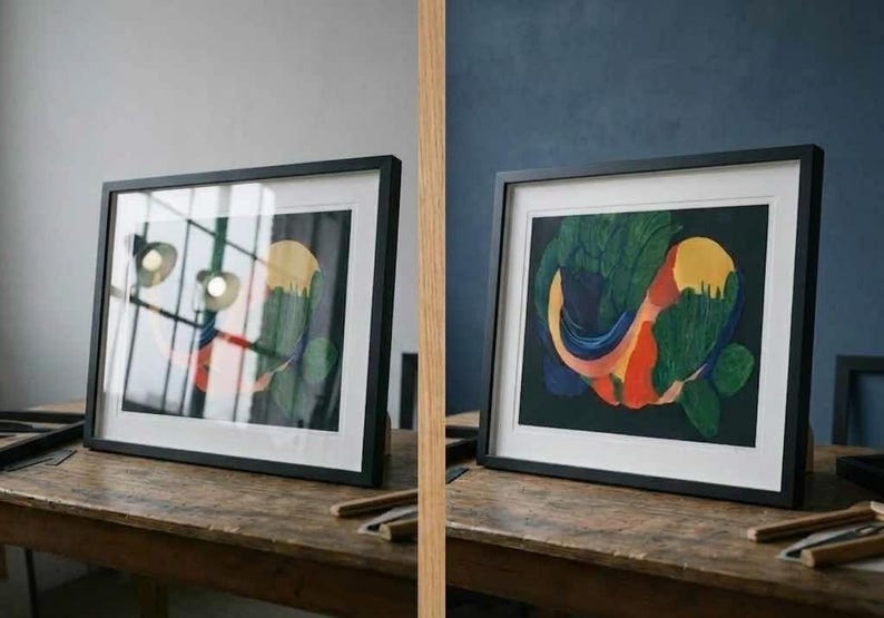 May include: Two framed abstract art prints. Each print features a colorful design with green, yellow, red, and blue shapes on a dark background. The frames are black and sit on a wooden table.