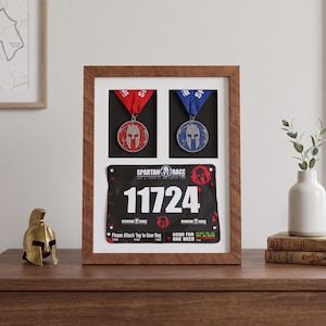 May include: A framed display featuring two Spartan Race medals with red and blue ribbons, and a race bib with the number 11724. The frame is brown and sits on a wooden surface. A gold helmet statue is on the left.