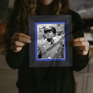 May include: A framed black and white photograph featuring a man in a police uniform. The photograph is set within a black frame with a blue mat. The man is wearing a hat and a double-breasted coat.