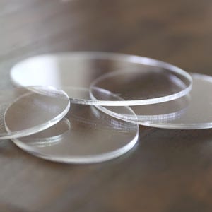 May include: A collection of clear, circular acrylic discs of varying sizes, scattered on a wooden surface. The transparent circles are stacked and overlapping, creating depth and dimension. The discs are smooth and reflective.
