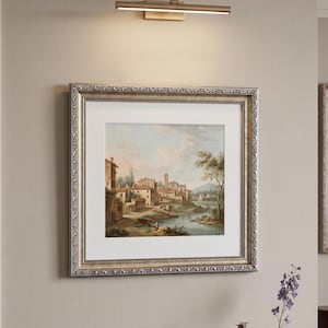 May include: A framed landscape painting with a silver ornate frame hangs on a beige wall. The painting depicts a European village scene with a river and buildings. A bronze-colored picture light is mounted above the artwork.