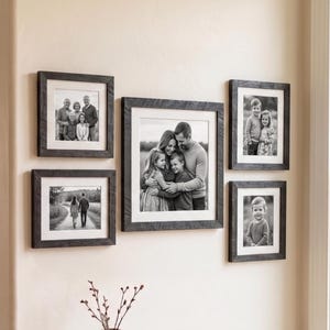 May include: A collection of five framed black and white photographs of family portraits and scenic views. The frames are dark gray with a textured appearance, mounted on a light beige wall. The photographs depict various family members and outdoor scenes.