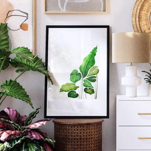 May include: A framed botanical print featuring a watercolor illustration of green leaves in various shapes and sizes. The artwork is set against a white background and framed in a black border. The print is displayed on a wooden side table.
