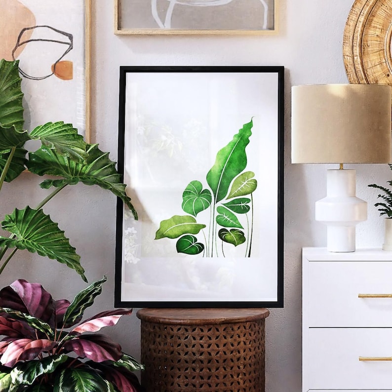 May include: A framed botanical print featuring a watercolor illustration of green leaves in various shapes and sizes. The artwork is set against a white background and framed in a black border. The print is displayed on a wooden side table.