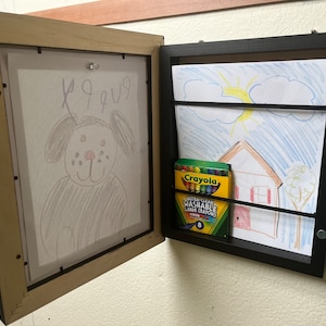 May include: A wall-mounted display frame with a hinged door, showcasing children's artwork. One side features a drawing of a dog, while the other displays a house and sun drawing, along with a box of crayons. The frame is black and beige.