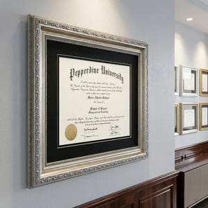 May include: A framed Pepperdine University diploma, featuring a silver ornate frame and a black mat. The diploma is displayed on a gray wall, with other framed documents visible in the background. The diploma is for a Master of Science in Management and Leadership.