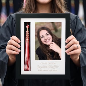 May include: A framed graduation photo featuring a smiling young person. The frame includes a tassel with "2024" and the graduate's name, "Jayden Shaffer," and school name. The frame is held by a person wearing a graduation gown.