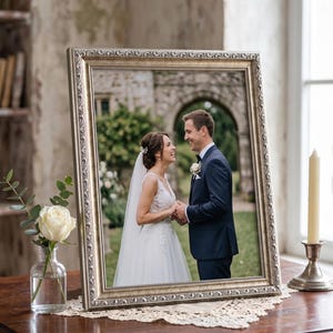 Op de afbeelding: Een ingelijste trouwfoto met een zilveren, sierlijke lijst. De foto toont een lachend stel dat elkaars handen vasthoudt. Een witte roos in een glazen vaas en een brandende kaars in een zilveren houder staan op de tafel.