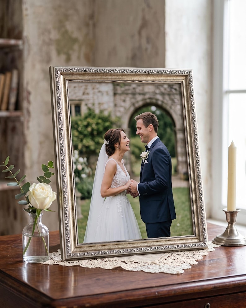Op de afbeelding: Een ingelijste trouwfoto met een zilveren, sierlijke lijst. De foto toont een lachend stel dat elkaars handen vasthoudt. Een witte roos in een glazen vaas en een brandende kaars in een zilveren houder staan op de tafel.