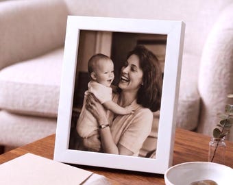 Può includere: Una cornice rettangolare bianca con una fotografia in seppia di una donna che tiene in braccio un bambino. La donna e il bambino sorridono. La cornice è su un tavolo di legno, con una piccola ciotola bianca e una pila di carta nelle vicinanze.