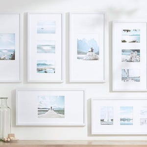 May include: A gallery wall of white picture frames with different landscape photos. The frames are arranged in a staggered pattern on a white wall.