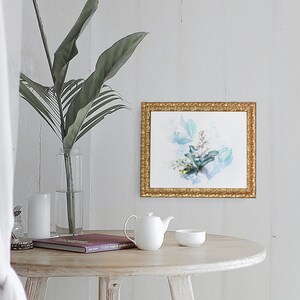 May include: A white ceramic teapot and a white ceramic bowl sit on a light wood table with a book and a vase of green leaves. A framed watercolor painting of blue and green leaves hangs on a white wall behind the table.