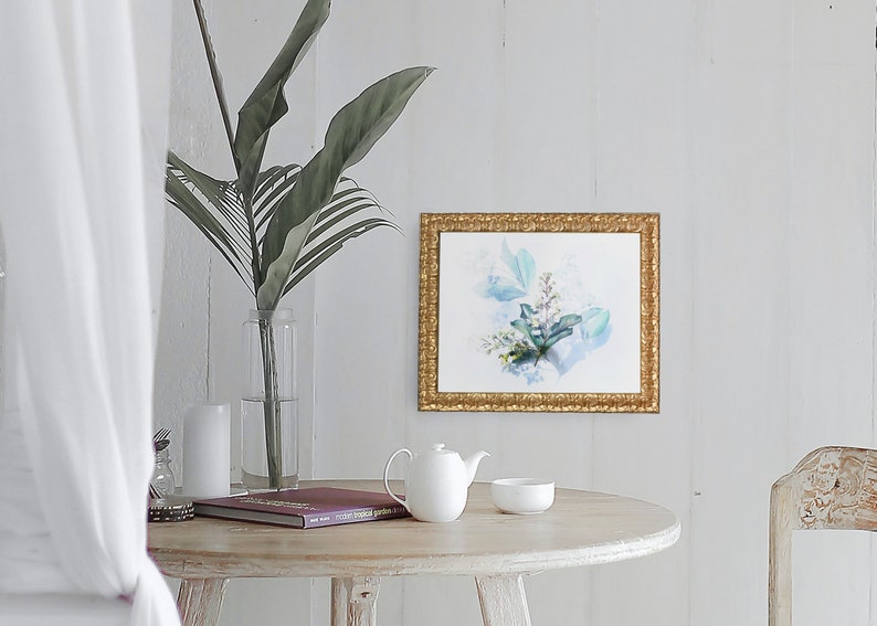 May include: A white ceramic teapot and a white ceramic bowl sit on a light wood table with a book and a vase of green leaves. A framed watercolor painting of blue and green leaves hangs on a white wall behind the table.