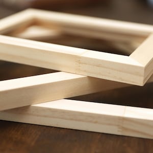 May include: Close-up of three unfinished wooden picture frames. The frames are rectangular with mitered corners, stacked on top of each other. The wood has a light, natural color, and the grain is visible. The frames are ready for artwork.