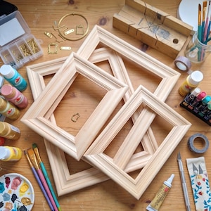 May include: A collection of unfinished wooden picture frames, art supplies, and tools arranged on a wooden surface. The items include paintbrushes, paint, a miter box, and various hardware pieces. The frames are rectangular with a decorative edge.