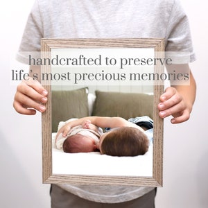Puede incluir: Una fotograf&iacute;a enmarcada de dos hermanos, un ni&ntilde;o mayor sosteniendo a un beb&eacute; m&aacute;s peque&ntilde;o. La foto est&aacute; en un marco de madera clara con un paspart&uacute; blanco. El texto "handcrafted to preserve life's most precious memories" est&aacute; impreso sobre el marco.
