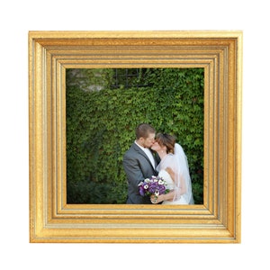 May include: A gold-colored picture frame with a wedding photo of a couple kissing in front of a green ivy wall. The bride is wearing a white dress and veil, and the groom is wearing a gray suit.