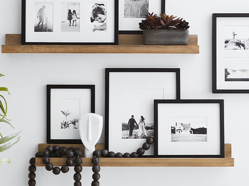 May include: A display of framed black and white photographs on wooden shelves. The frames vary in size and contain images of people, landscapes, and abstract designs. A decorative wooden bead garland and a small succulent plant are also present.
