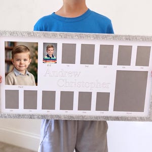 May include: A framed school photo display with a distressed white finish. The display includes spaces for photos from K-5 and beyond, with the name "Andrew Christopher" in the center. The frame is held by a person.
