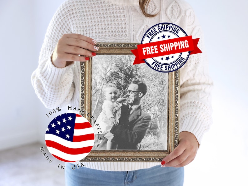 Op de afbeelding: Een ingelijste zwart-witfoto van een man die een baby vasthoudt. De lijst is goudkleurig met sierlijke details. De afbeelding is gelabeld met "100% Handcrafted Made in USA".