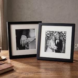 May include: Two black framed wedding photos with white matting sit on a wooden table. One shows hands with rings, the other a couple. A candle in a brass holder and a small vase are also on the table.