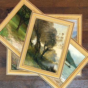 May include: Four framed landscape paintings with gold-colored frames are arranged on a dark wood surface. The paintings depict trees, water, and grassy areas, with a muted color palette of greens, browns, and blues.