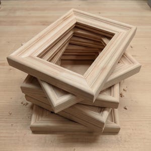 May include: A stack of unfinished wooden picture frames. The frames are rectangular with a beveled edge and a recessed inner area. The wood has a light, natural color and visible grain.