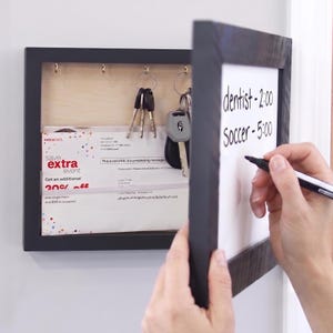 May include: A black wall-mounted organizer with a hinged whiteboard front featuring the handwritten text dentist-2:00 and Soccer-5:00. The interior contains metal hooks holding keys and a slot for mail, with a light wood back panel.
