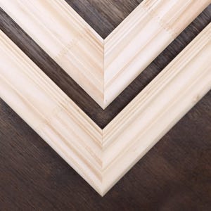 May include: Close-up of three unfinished wooden picture frames arranged in a V-shape. The frames are made of light-colored wood, with a natural grain pattern. The background is a dark brown wooden surface.