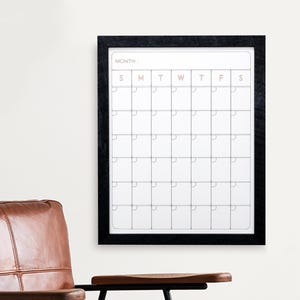 May include: A black framed monthly calendar with a white interior, hanging on a white wall. The calendar grid is divided into squares for each day of the month. The word "MONTH" is at the top.