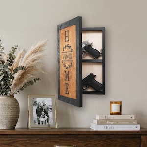May include: A wooden wall-mounted gun safe with a black frame and a hinged door. The door features the word "HOME" and the family name "Vitelli". Two black handguns are visible inside the safe. The safe is mounted on a beige wall.