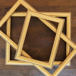 May include: Overhead shot of three gold picture frames arranged on a dark brown wooden surface. The frames are rectangular with a decorative border and a gold finish. The frames are empty, and the composition is simple.