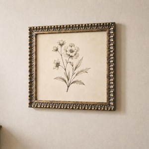 Op de afbeelding: Een vierkant ingelijst kunstwerk aan de muur met een zwart-witte botanische schets van een bloem met bladeren op een crèmekleurige achtergrond. De lijst is gemaakt van donker, getextureerd bronskleurig metaal met een schulprandpatroon.