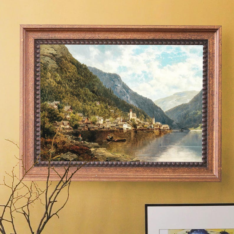 May include: A framed landscape painting depicts a lakeside village nestled at the base of towering mountains. The artwork features a serene lake reflecting the sky and surrounding buildings. The frame is a warm, light brown with a beaded edge.