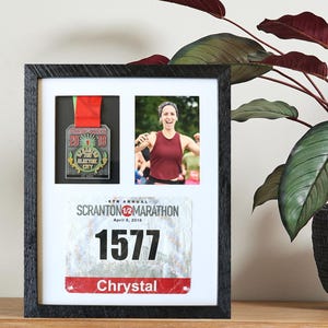 May include: A framed display featuring a marathon medal, a photo of a runner, and a race bib. The medal has a red ribbon and the text "The Electric City". The race bib displays the number 1577 and the name "Chrystal".