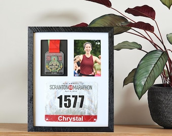 May include: A framed display featuring a marathon medal, a photo of a runner, and a race bib. The medal has a red ribbon and the text "The Electric City". The race bib displays the number 1577 and the name "Chrystal".