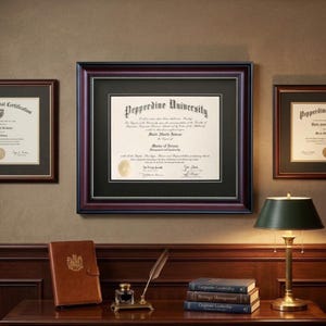 May include: Framed Pepperdine University diploma with a black and burgundy frame, displayed on a wall with other framed documents. A desk with a leather journal, books, a lamp, and an inkwell is in the foreground.