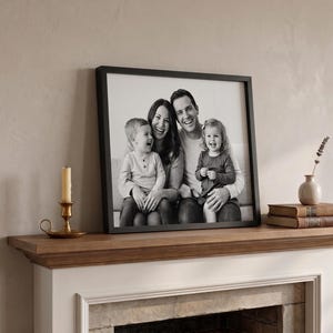 May include: A framed black and white family portrait featuring a smiling couple and their two young children. The photograph is displayed on a wooden mantelpiece, alongside a candle, a vase, and books.