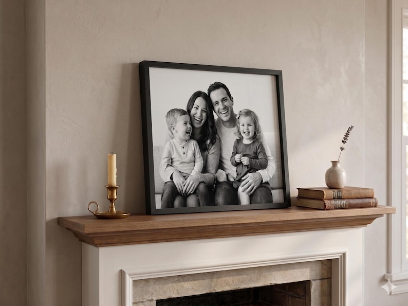 May include: A framed black and white family portrait featuring a smiling couple and their two young children. The photograph is displayed on a wooden mantelpiece, alongside a candle, a vase, and books.