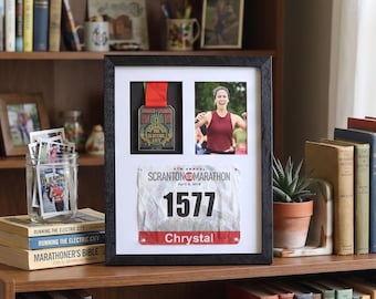 Può includere: Un espositore incorniciato nero con una medaglia da maratona, una foto di una corridora e un pettorale. La medaglia è rossa e dorata. Il pettorale recita "SCRANTON MARATHON" e "1577 Chrystal". È posto su una mensola di legno.