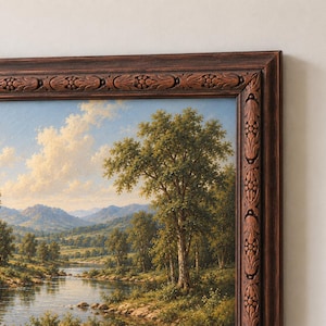 Op de afbeelding: Een landschapsolieverfschilderij in een donkerbruine houten lijst met uitgesneden bloemendetails. Het kunstwerk toont een rivier die door een groene vallei stroomt met bomen, glooiende heuvels en verre bergen onder een blauwe lucht.