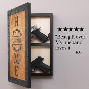 May include: A wooden wall-mounted gun cabinet with a black frame and a light brown interior. The cabinet door has the word "HOME" and the name "Vitelli" engraved on it. Two handguns are visible inside the cabinet.